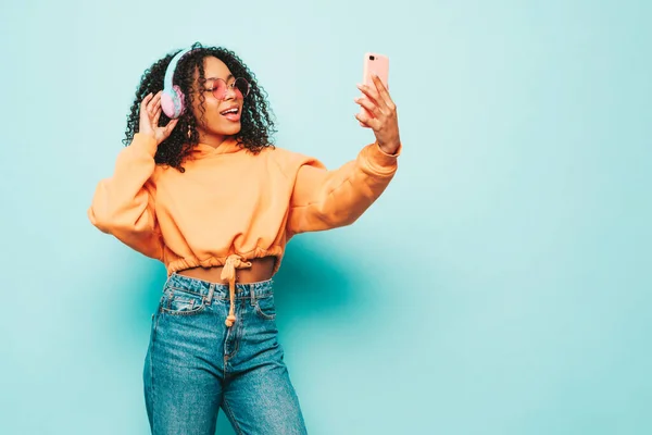 Beautiful black woman with afro curls hairstyle.Smiling model in orange hoodie and jeans.Sexy carefree female listening music in wireless headphones.Posing in studio near blue wall.Taking selfie