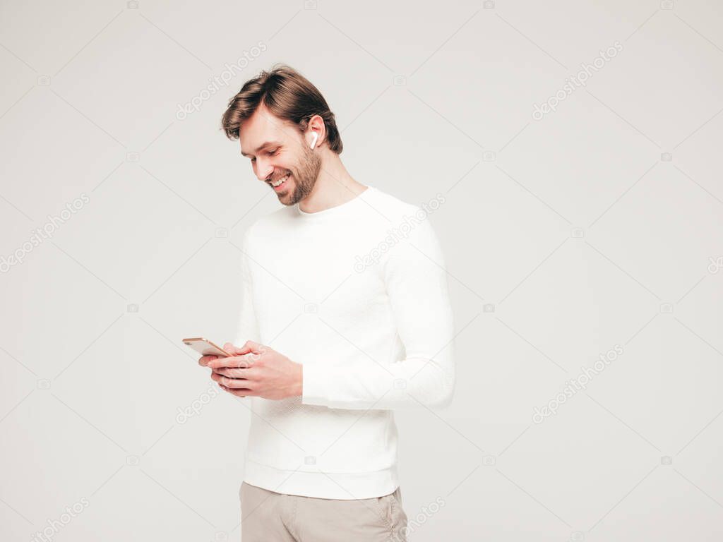 Handsome smiling hipster lumbersexual businessman model wearing white casual sweater and trousers.Fashion stylish man posing against gray background in studio. Holding smartphone in hands.In earphones