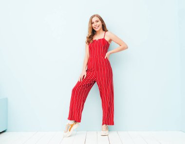 Yeni moda kırmızı takım elbiseli güzel, gülümseyen bir kadının portresi. Stüdyoda açık mavi duvarın yanında poz veren kaygısız seksi kadın. Pozitif model içeride eğleniyor. Neşeli ve mutlu