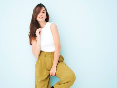 Moda kıyafetleri içinde gülümseyen genç ve güzel bir bayan. Stüdyoda açık mavi duvarın yanında poz veren kaygısız seksi kadın. Pozitif model içeride eğleniyor. Neşeli ve mutlu