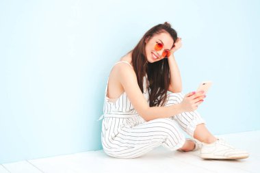 Son moda yaz hippileri içinde gülümseyen genç bir bayan tulum giyiyor. Stüdyoda açık mavi duvarın yanında poz veren kaygısız seksi kadın. Pozitif model içeride eğleniyor. Akıllı telefon uygulamaları kullanmaktan keyif alıyorum.