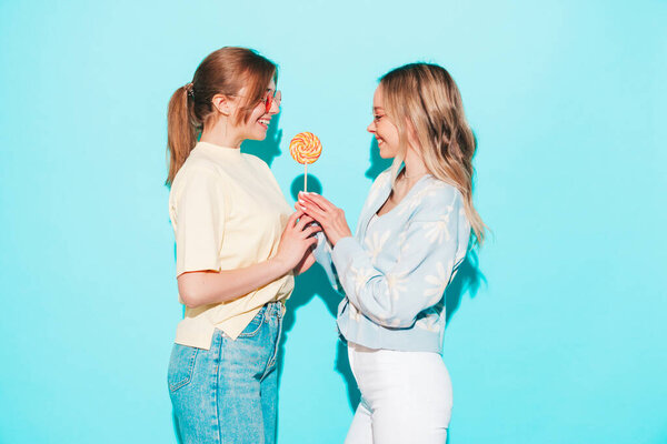 Two young beautiful blond smiling hipster female in trendy summer clothes. Sexy carefree women posing near blue wall in studio. Trendy and positive models having fun with candy sweet lollipop