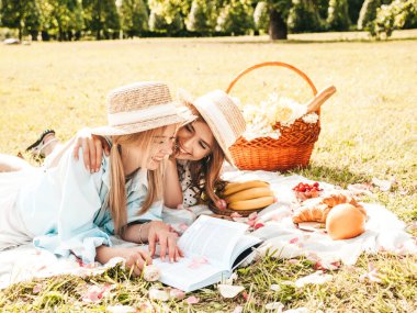 Moda elbisesi ve şapkası içinde iki güzel bayan. Dışarıda piknik yapan kaygısız kadınlar. Çimlerin üzerinde oturmuş meyve ve peynir yiyen pozitif modeller.