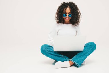 Saçları kıvırcık, güzel siyah bir kadın. Süveter ve kotlar içinde gülümseyen bir model. Stüdyoda beyaz arka planda oturan kaygısız seksi kadın. Bronz tenli ve neşeli. Laptopunu kullanıyor.