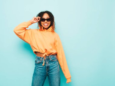 Afro kıvırcık saçlı, turuncu kapüşonlu ve kotlu güzel siyah kadın. Stüdyoda güneş gözlükleriyle mavi duvarın yanında poz veren kaygısız seksi kadın.