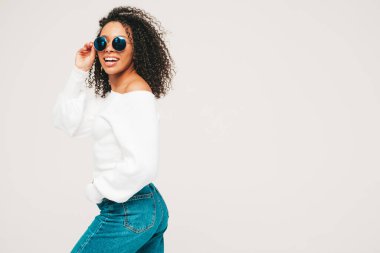 Saçları kıvırcık, güzel siyah bir kadın. Süveter ve kotlar içinde gülümseyen bir model. Stüdyoda beyaz arka planda poz veren kaygısız seksi kadın. Bronzlaşmış ve neşeli. Güneş gözlükleriyle
