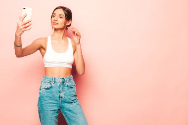 Beyaz formalı bluz ve kot pantolon giymiş güzel bir kadın. Seksi, kaygısız, neşeli manken içeride eğleniyor. Stüdyoda pembe duvarın yanında poz veren sevimli ve pozitif kadın. Selfie çekiyor. 