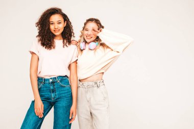 Moda kıyafetleri içinde iki güzel, gülümseyen uluslararası hippi kadın. Stüdyoda gri arka planda poz veren kaygısız kadınlar. Güneş gözlükleriyle eğlenen pozitif modeller.