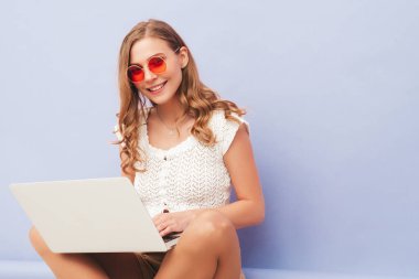 Son moda yaz hippileri içinde gülümseyen genç bir bayan tulum giyiyor. Stüdyoda açık mor duvarın yanında poz veren kaygısız seksi kadın. Pozitif model içeride eğleniyor. Bilgisayarını kullanıyor.