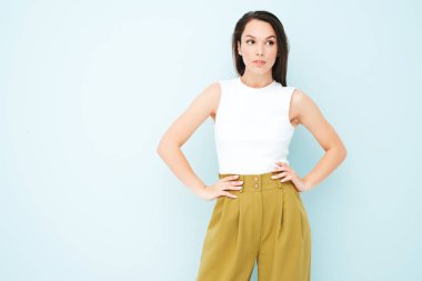 Moda kıyafetleri içinde gülümseyen genç ve güzel bir bayan. Stüdyoda açık mavi duvarın yanında poz veren kaygısız seksi kadın. Pozitif model içeride eğleniyor. Neşeli ve mutlu