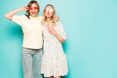 Moda kıyafetleri içinde gülümseyen iki güzel sarışın hippi kadın. Stüdyoda mavi duvarın yanında poz veren kaygısız seksi kadınlar. Moda ve pozitif modeller eğleniyor.