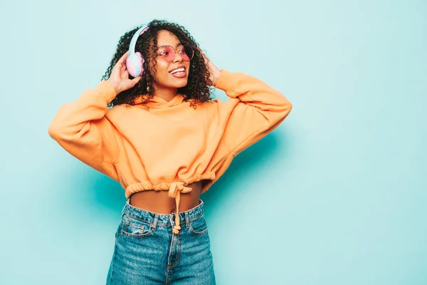 Beautiful black woman with afro curls hairstyle.Smiling model in orange hoodie and jeans.Sexy carefree female listening music in wireless headphones.Posing in studio near blue wall