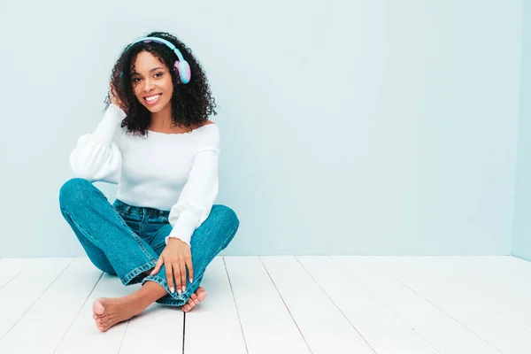 Beautiful black woman with afro curls hairstyle.Smiling model in sweater and jeans.Sexy carefree female listening music in wireless headphones.Posing in studio near light blue wall