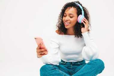 Saçları kıvırcık, saçları kıvırcık, süveter ve kot giyen, kablosuz kulaklıkla müzik dinleyen kaygısız bir kadın. Stüdyoda beyaz arka planda poz veriyor. Akıllı telefon tutuyor.