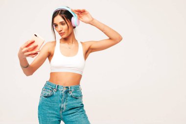 Kot pantolon giymiş, gülümseyen güzel bir kadın. Kablosuz kulaklıkla müzik dinleyen seksi tasasız model. Stüdyoda gri arka planda poz veren sevimli ve pozitif kadın. Selfie çekiyor.