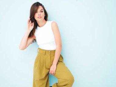 Moda kıyafetleri içinde gülümseyen genç ve güzel bir bayan. Stüdyoda açık mavi duvarın yanında poz veren kaygısız seksi kadın. Pozitif model içeride eğleniyor. Neşeli ve mutlu