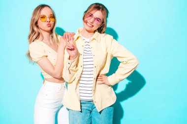 Moda kıyafetleri içinde gülümseyen iki güzel sarışın hippi kadın. Stüdyoda mavi duvarın yanında poz veren kaygısız seksi kadınlar. Moda ve pozitif modeller güneş gözlükleriyle eğleniyor.