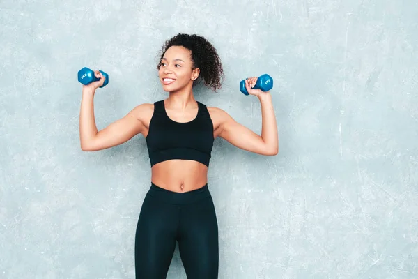 Fitness smiling black woman in sports clothing with afro curls hairstyle.She wearing sportswear. Young beautiful model with perfect tanned body.Female holding dumbbells in studio near grey wall