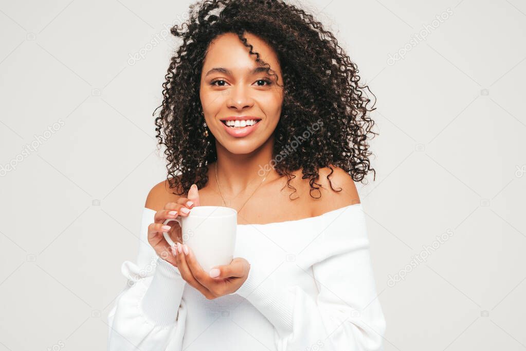 Beautiful black woman with afro curls hairstyle.Smiling model in sweater and trendy jeans clothes. Sexy carefree female posing on white background in studio. Drinking coffee. Holding cup in hands