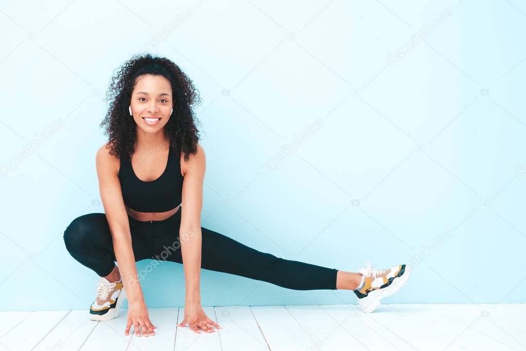 Portrait of fitness smiling black woman in sports clothing with afro curls hairstyle.She wearing sportswear.Young beautiful model stretching out before training.Female sitting in studio near blue wall