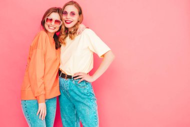 İki genç, güzel, gülümseyen yeniyetme kadın, modaya uygun yaz hippi kıyafetleri içinde. Stüdyoda pembe duvarın yanında poz veren kaygısız seksi kadınlar. Moda ve pozitif modeller eğleniyor. Neşeli ve mutlu