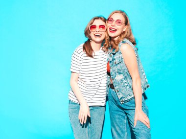 Moda kıyafetleri içinde gülümseyen iki güzel hippi kadın. Stüdyoda mavi duvarın yanında poz veren kaygısız seksi kadınlar. Moda ve pozitif modeller eğleniyor. Neşeli ve mutlu