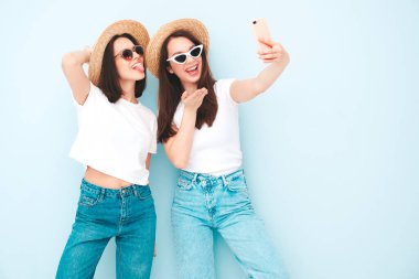 Son moda beyaz tişörtlü ve kot pantolonlu iki güzel gülümseyen hippi kadın. Stüdyoda açık mavi duvarın yanında poz veren kaygısız kadınlar. Şapkalı ve şapkalı pozitifler.