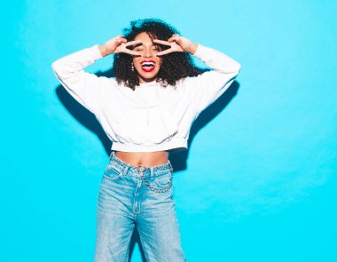 Saçları kıvırcık, güzel siyah bir kadın. Moda kıyafetleri içinde gülümseyen bir model. Stüdyoda mavi duvarın yanında poz veren kaygısız seksi kadın. Kırmızı rujlarla bronzlaşmış ve neşeli. Barış işareti nasıl?