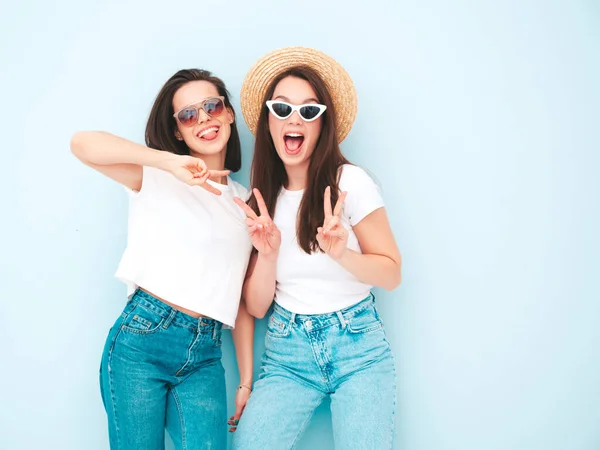 流行の夏の白いTシャツとジーンズの服で2人の若い美しい笑顔のヒップスター女性 スタジオでライトブルーの壁の近くにポーズセクシーな屈託のない女性 帽子の肯定的なモデル 平和のサインを表示 — ストック写真