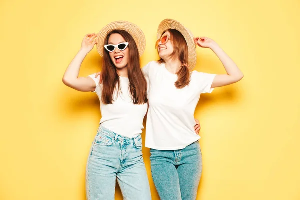 流行の夏の白いTシャツとジーンズの服で2人の若い美しい笑顔ヒップスター女性 スタジオで黄色の壁の近くにポーズセクシーな屈託のない女性 帽子の肯定的なモデル パーティーで踊る — ストック写真