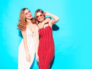 İki genç, güzel, gülümseyen hippi kadın, modaya uygun yaz tulumları giyiyor. Stüdyoda mavi duvarın yanında poz veren kaygısız seksi kadınlar. Moda ve pozitif modeller eğleniyor. Neşeli ve mutlu