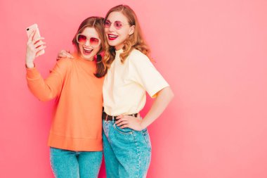 İki genç, güzel, gülümseyen yeniyetme kadın, modaya uygun yaz hippi kıyafetleri içinde. Stüdyoda pembe duvarın yanında poz veren kaygısız seksi kadınlar. Moda ve pozitif modeller eğleniyor. Selfie çekiyoruz.