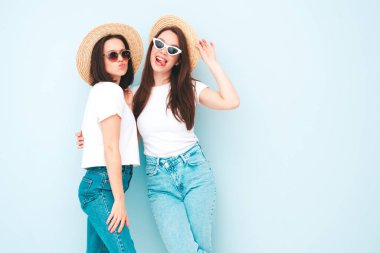 Son moda beyaz tişört ve kot pantolon giyen iki güzel, gülümseyen yeniyetme kadın. Stüdyoda açık mavi duvarın yanında poz veren kaygısız kadınlar. Şapkalı olumlu modeller.