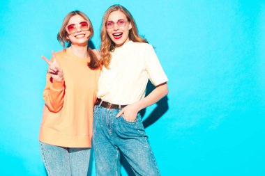 İki genç, güzel, gülümseyen yeniyetme kadın, modaya uygun yaz hippi kıyafetleri içinde. Stüdyoda mavi duvarın yanında poz veren kaygısız seksi kadınlar. Moda ve pozitif modeller eğleniyor. Neşeli ve mutlu