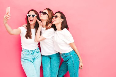 Aynı yaz moda olan beyaz tişört ve kot pantolon giyen üç güzel, gülümseyen hippi kadın. Stüdyoda pembe duvarın yanında poz veren kaygısız seksi kadınlar. Güneş gözlüklü pozitifler selfie çekiyor.