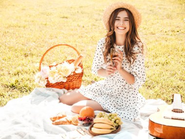 Moda elbisesi ve şapkası olan genç ve güzel hippi kadın. Kaygısız kadın dışarıda piknik yapıyor. Pozitif model çimlerin üzerinde oturuyor, meyve ve peynir yiyor, şampanya içiyor.