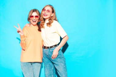 Moda kıyafetleri içinde gülümseyen iki güzel hippi kadın. Stüdyoda mavi duvarın yanında poz veren kaygısız seksi kadınlar. Moda ve pozitif modeller eğleniyor. Neşeli ve mutlu