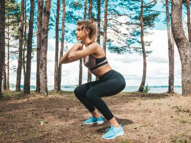 Parktaki konsantre olmuş genç kadın portresi. Model, antrenmandan önce spor esneme egzersizleri yapar. Ormanda kadın poz veriyor. Spor dersinde. Çömelme yapıyor. 