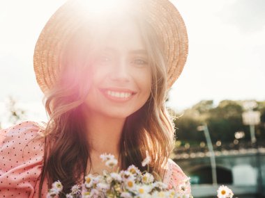 Genç, güzel, güler yüzlü yeniyetme kadın. Moda bir yaz elbisesi içinde. Günbatımında şapkayla sokakta poz veren kaygısız seksi kadın. Arazi seti üzerinde pozitif model. Çiçekleri açık havada tutmak