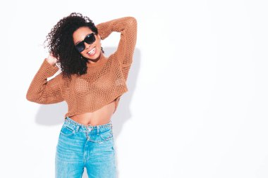 Saçları kıvırcık, güzel siyah bir kadın. Moda kıyafetleri içinde gülümseyen bir model. Stüdyodaki beyaz duvarda izole edilmiş kaygısız seksi kadın. Bronzlaşmış ve neşeli 