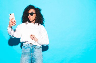 Saçları kıvırcık, güzel siyah bir kadın. Moda kıyafetleri içinde gülümseyen bir model. Stüdyoda mavi duvarın yanında poz veren kaygısız seksi kadın. Bronzlaşmış ve neşeliydi. Güneş gözlükleriyle selfie çekiyordu.