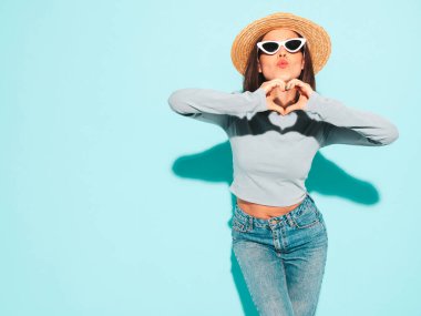 Son moda yaz hippi kıyafetleri içinde gülümseyen güzel bir kadının portresi. Stüdyoda mavi duvarın yanında poz veren kaygısız seksi kadın. Pozitif model güneş gözlüğü ve şapkayla eğleniyor. Kalp işareti yapar