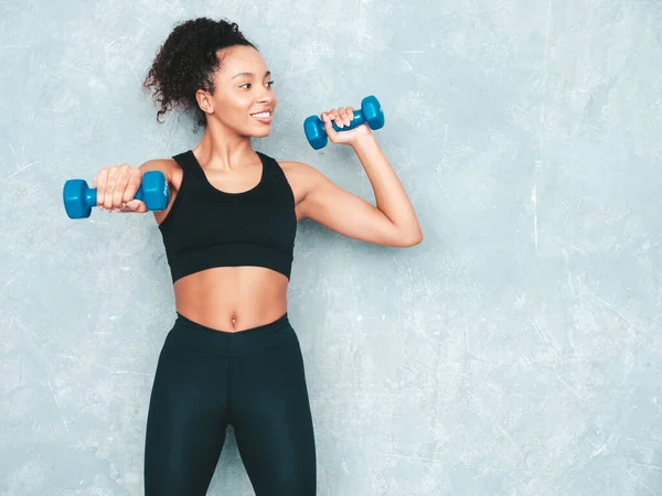 Fitness smiling black woman in sports clothing with afro curls hairstyle.She wearing sportswear. Young beautiful model with perfect tanned body.Female holding dumbbells in studio near grey wall