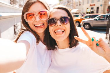 Son moda beyaz tişörtlü ve kot pantolonlu iki güzel, gülümseyen yeniyetme bayan sokak arka planında poz veren kaygısız kadınlar. Pozitif mankenler eğleniyor, sarılıyor. Pov selfie çekiyor.