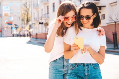 Son moda beyaz tişörtlü ve kot pantolonlu iki güzel gülümseyen yeniyetme kadın. Sokakta arka planda poz veren kaygısız kadınlar. Akıllı telefon uygulamaları kullanan, ekrana bakan pozitif modeller.