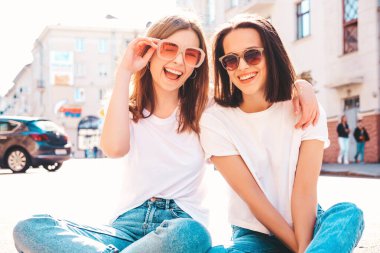Son moda beyaz tişörtlü ve kot pantolonlu iki güzel, gülümseyen hippi kadın. Sokakta arka planda oturan kaygısız kadınlar. Pozitif modeller eğleniyor, sarılıyor ve deliriyor.