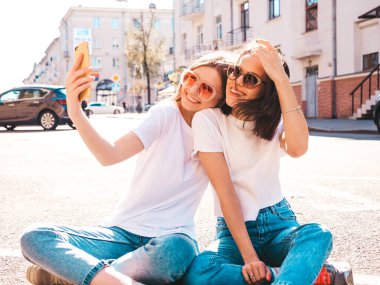 Son moda beyaz tişörtlü ve kot pantolonlu iki güzel, gülümseyen hippi kadın. Sokakta arka planda oturan kaygısız kadınlar. Pozitif modeller eğleniyor, selfie çekiyor.