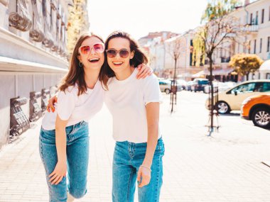 Son moda beyaz tişörtlü ve kot pantolonlu iki güzel, gülümseyen yeniyetme bayan sokak arka planında poz veren kaygısız kadınlar. Pozitif modeller eğleniyor, sarılıyor ve deliriyor.