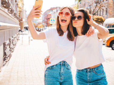 Son moda beyaz tişörtlü ve kot pantolonlu iki güzel gülümseyen yeniyetme kadın. Arka planda poz veren kaygısız kadınlar. Pozitif mankenler eğleniyor, sarılıyor.