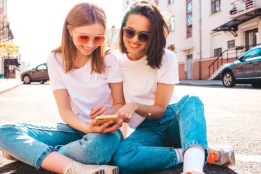 Son moda beyaz tişörtlü ve kot pantolonlu iki güzel gülümseyen yeniyetme kadın. Sokakta arka planda poz veren kaygısız kadınlar. Akıllı telefon uygulamaları kullanan, ekrana bakan pozitif modeller.